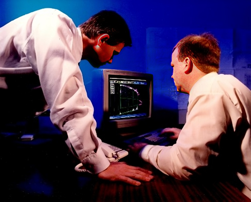 Two men in a room, both looking at a computer screen showing technical software.